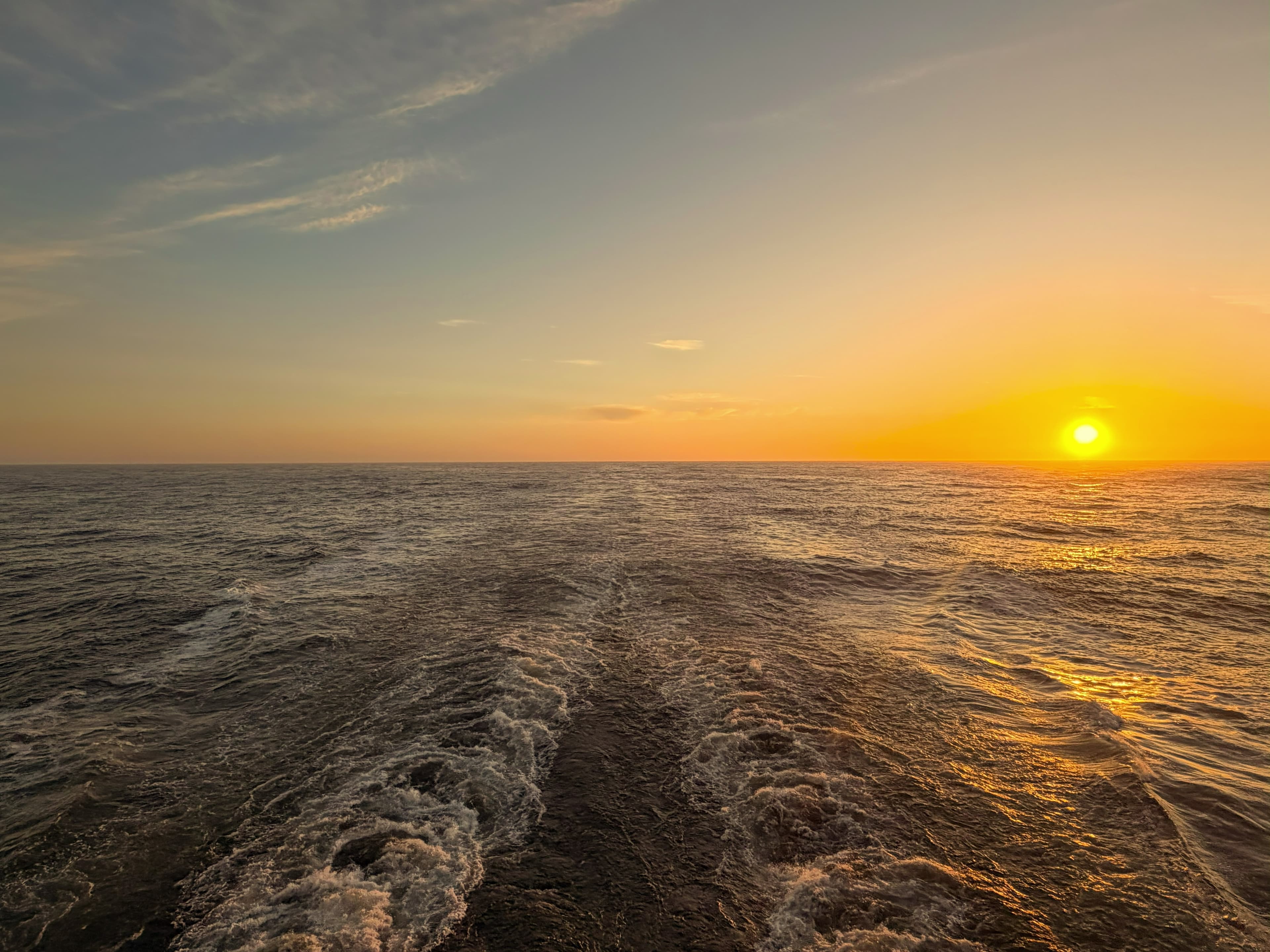 Sunset over open ocean, seen across a cruise ship's wake
